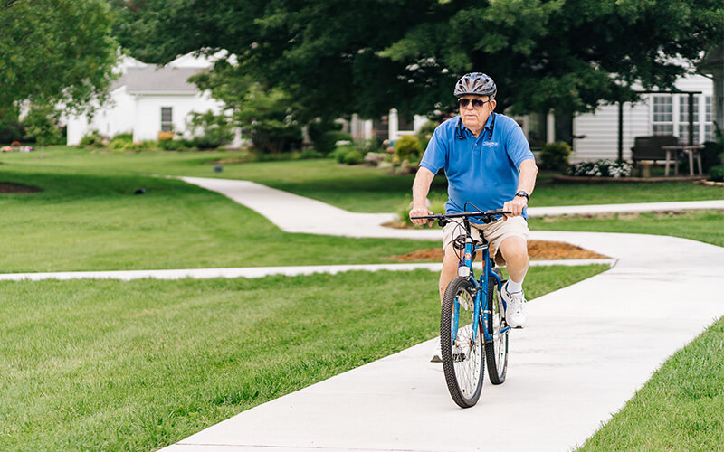 Senior man in blue shirt biking on a path near residential units.