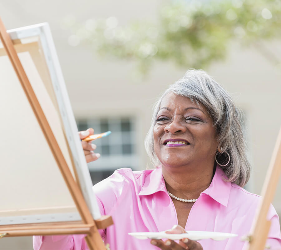 Smiling older woman in pink painting outdoors at a senior living community.