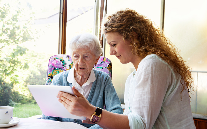 Young woman assists elderly woman with a tablet in a bright room.