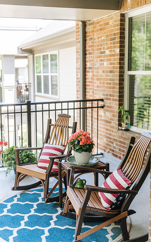 Outdoor balcony with two rocking chairs, flag-themed cushions, and potted flowers.