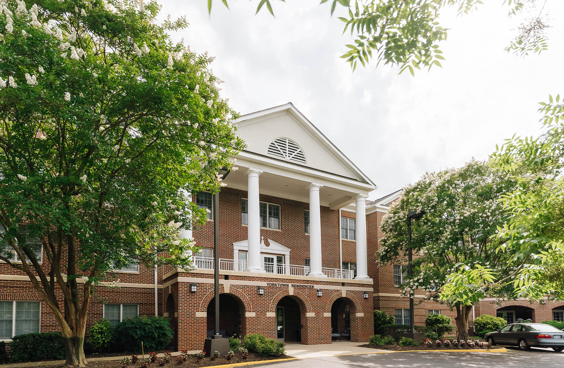 Brick building entrance with white columns and lush trees around parking area.