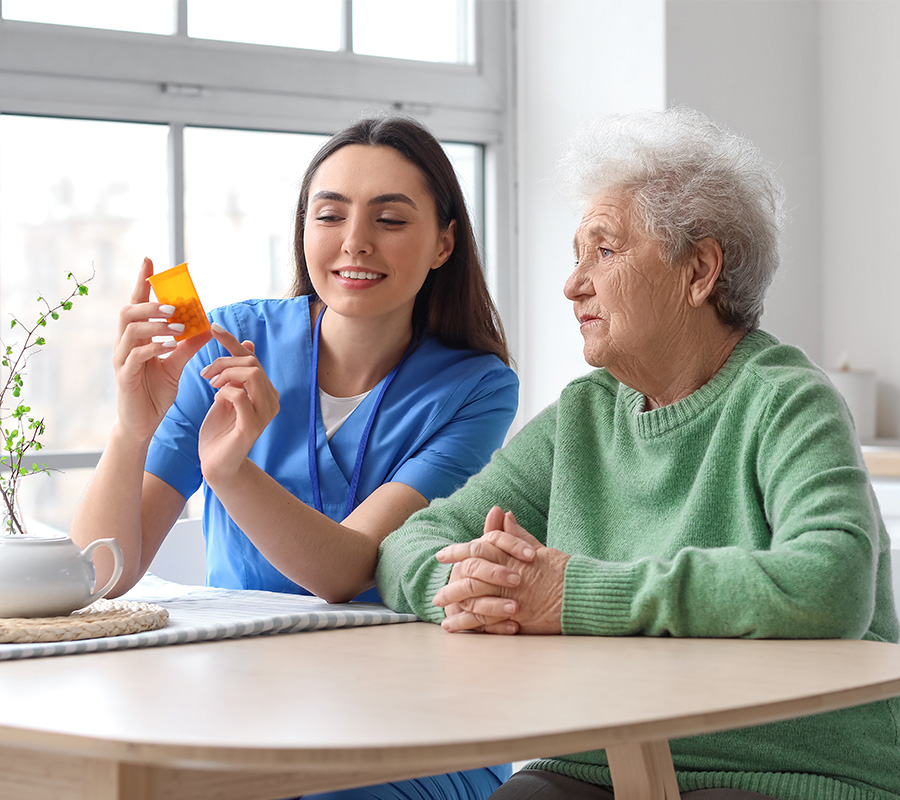 Caregiver in blue scrubs discusses medication with elderly woman in a bright living space.