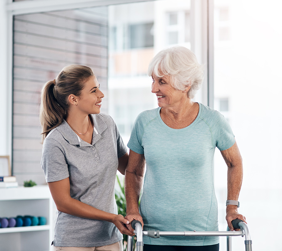 Elderly woman using walker, assisted by caregiver in a bright room.