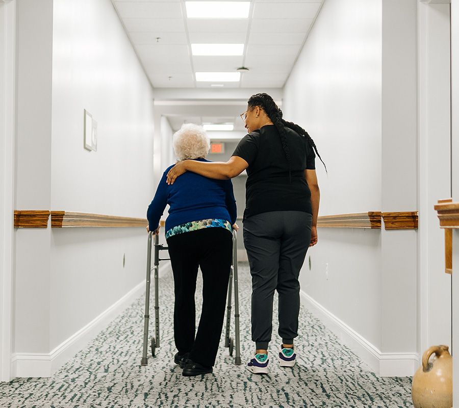 Caregiver assists senior woman with walker in a well-lit hallway.