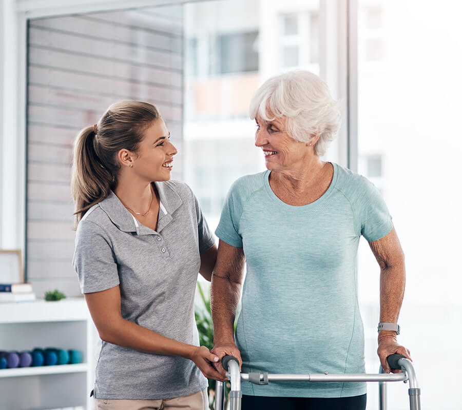 Caregiver assisting senior woman with walker indoors, both smiling.