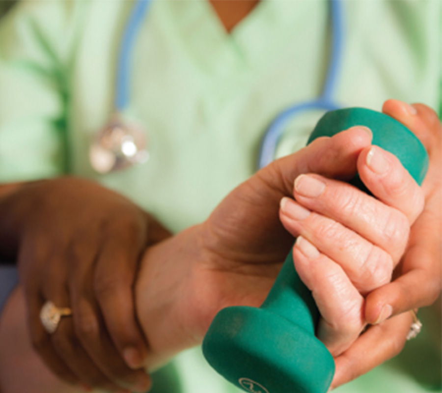 A caregiver assists a senior with a green dumbbell exercise.