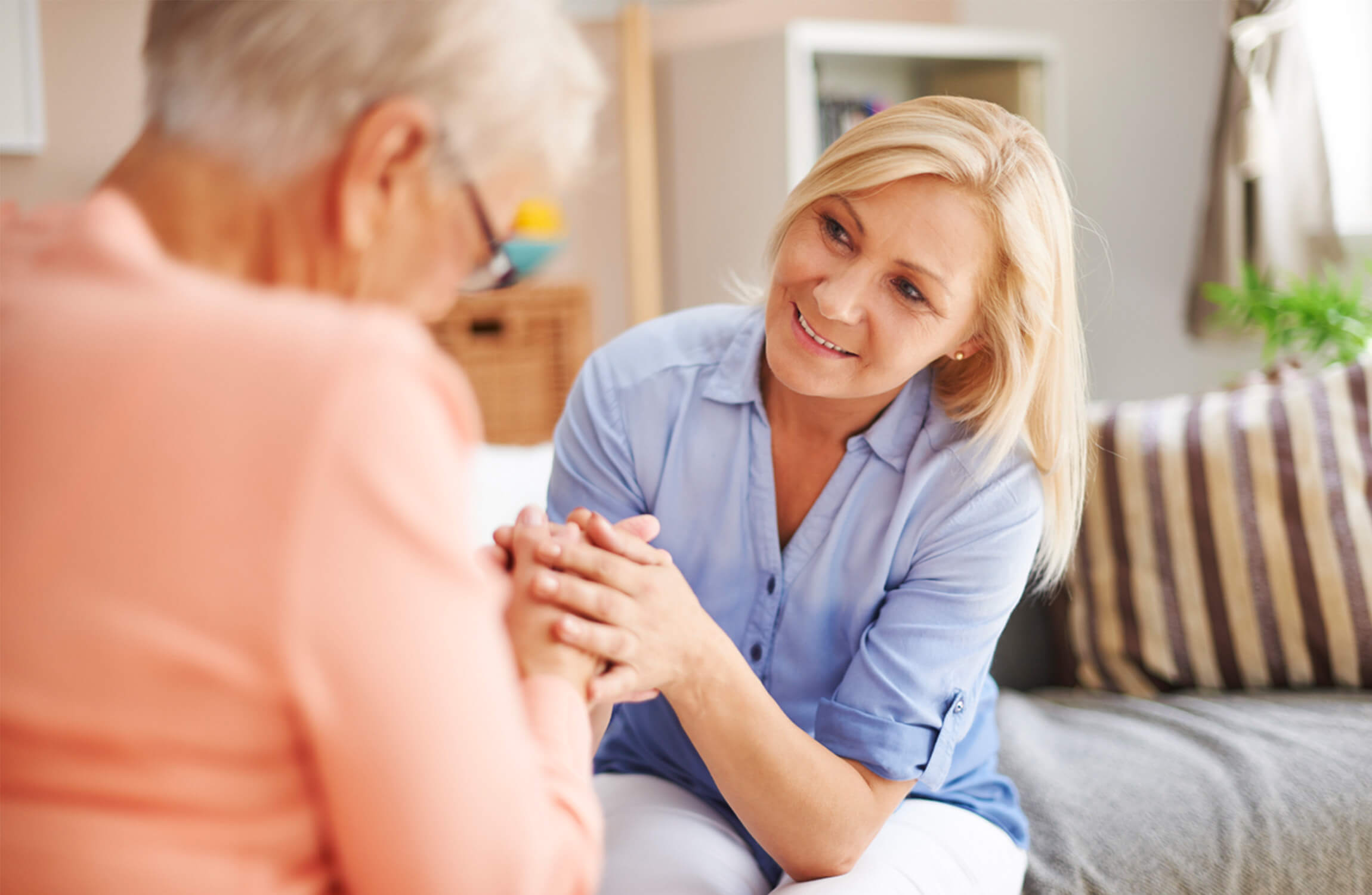 Caregiver smiling while holding hands with an older adult in a cozy room.