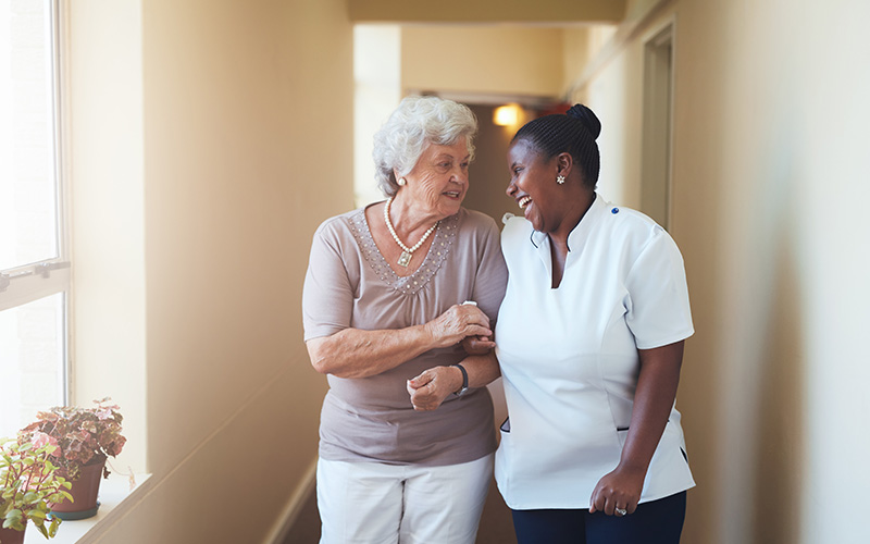 Caregiver and senior woman walking together in a warmly lit hallway.