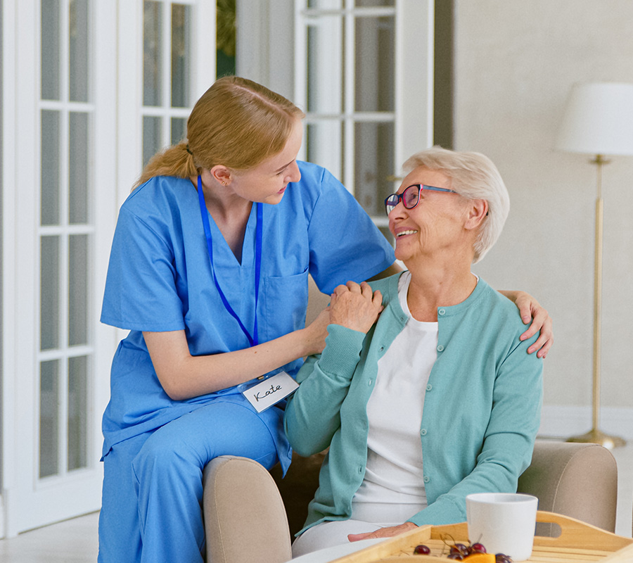 Caregiver supporting a smiling elderly woman in a bright living space, showing care and compassion.