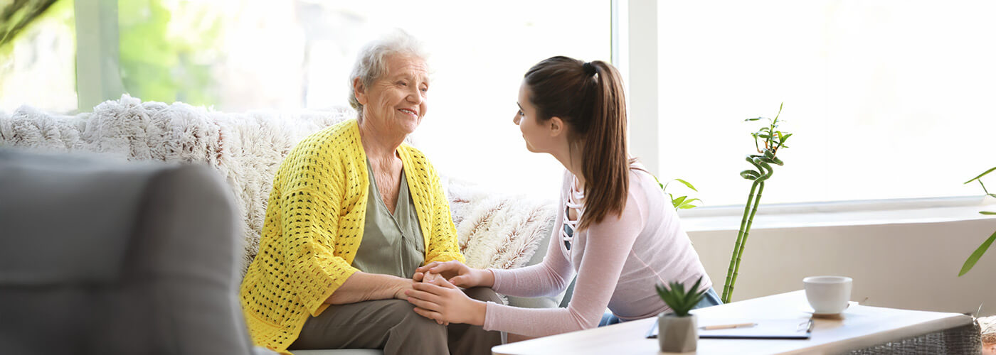Young woman converses with elderly woman on a cozy sofa in a bright, sunny room.