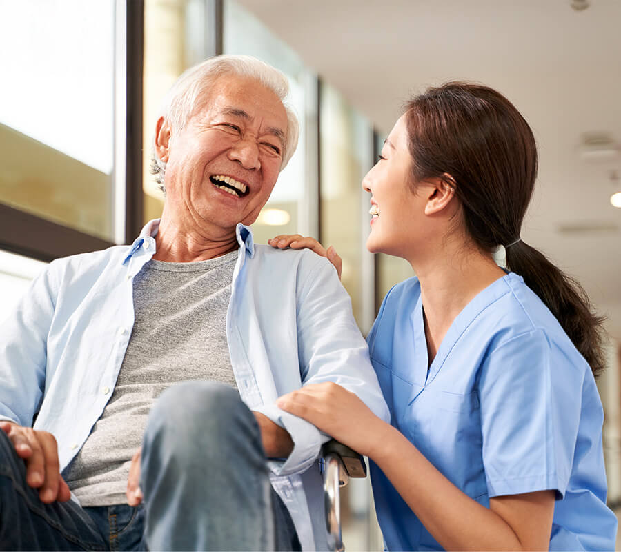 Elderly man in wheelchair smiling with nurse in light-filled corridor.