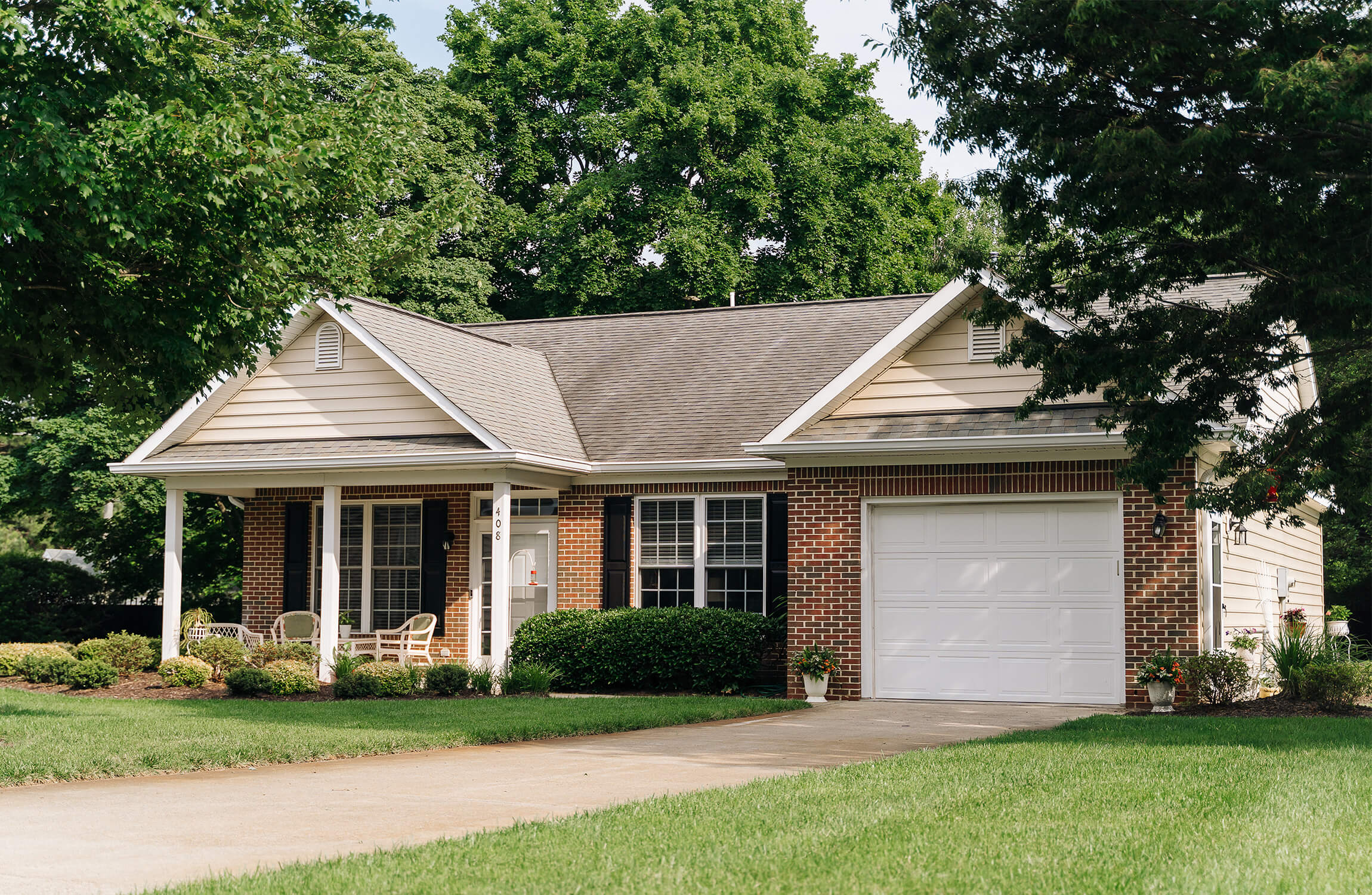 Charming Brick Unit Amidst Greenery Brick unit with white garage surrounded by lush green trees and lawn.