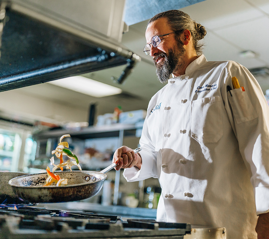 Chef in white coat tossing vegetables in pan in a professional kitchen.