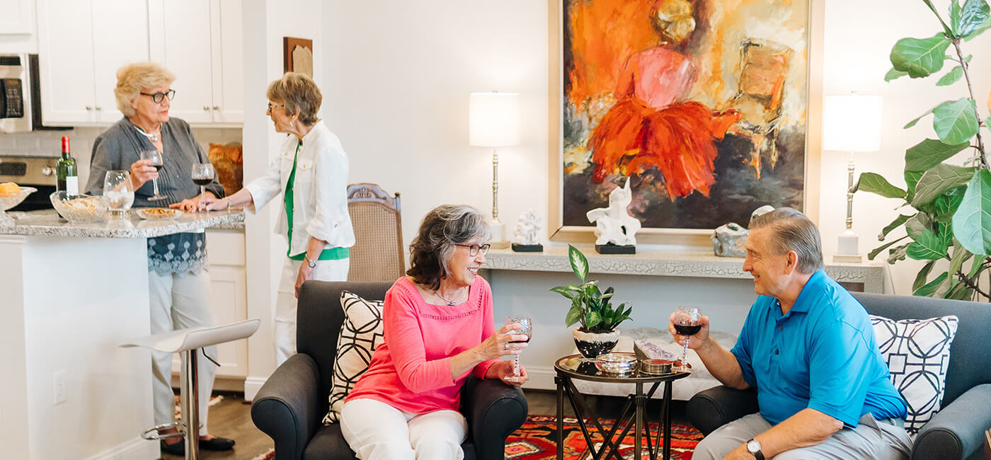 Four seniors enjoying wine in a cozy living room and kitchen area of a communal unit.