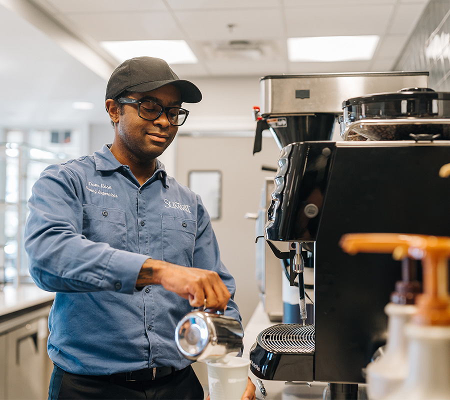 Staff member in blue shirt prepares coffee in community dining area.