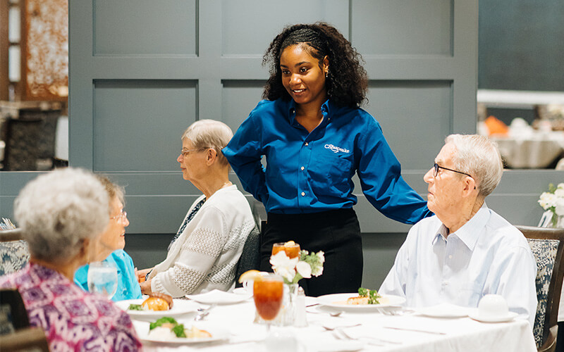 Dining area of a community with residents and a server engaged in conversation.