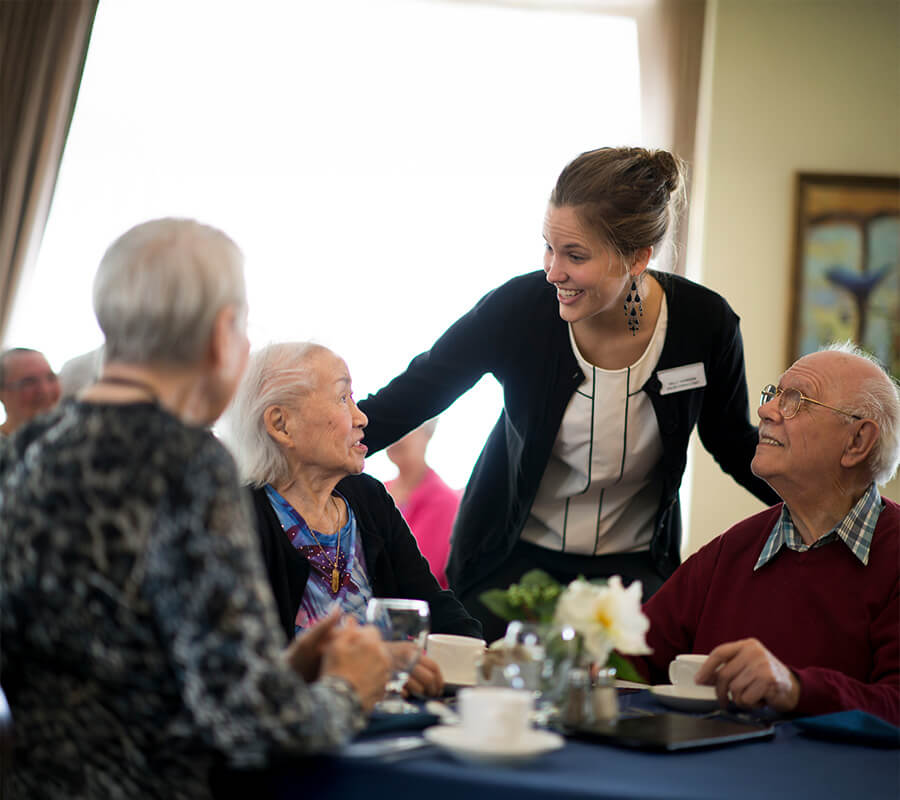 Staff member engages with residents at a dining table in a friendly atmosphere.