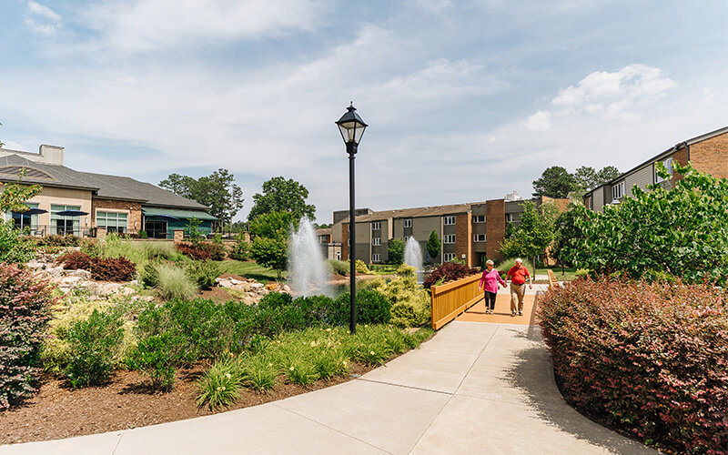 Peaceful Walk in the Community Garden Two people walking on a path in a landscaped garden with fountains and units in the background.