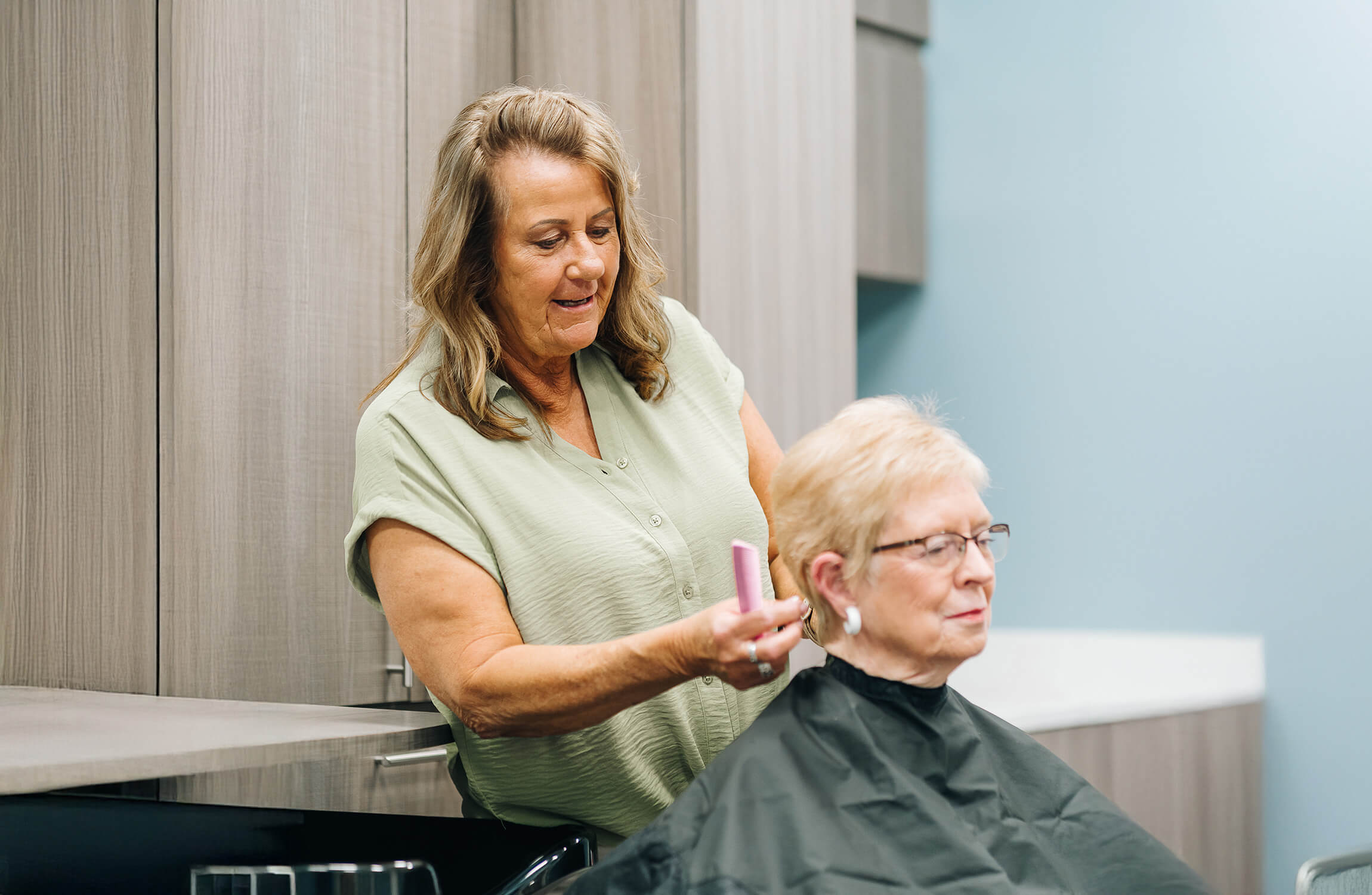 A woman in a green shirt cuts the hair of a seated person in a salon setting.