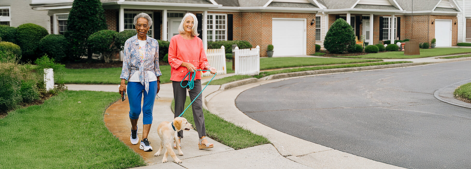 Two women walking a dog outside residential units in a green, landscaped community.