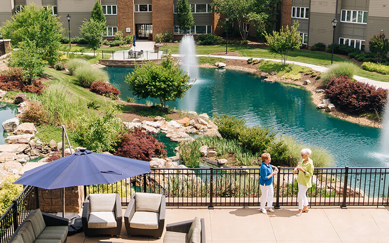 Courtyard with Pond and Seating Area Two people talking by a pond in a senior living community courtyard.