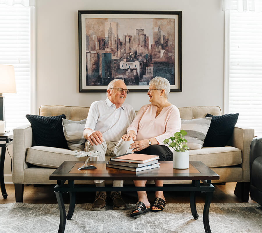 Older couple sitting on a sofa in a cozy living room with a cityscape painting behind them.