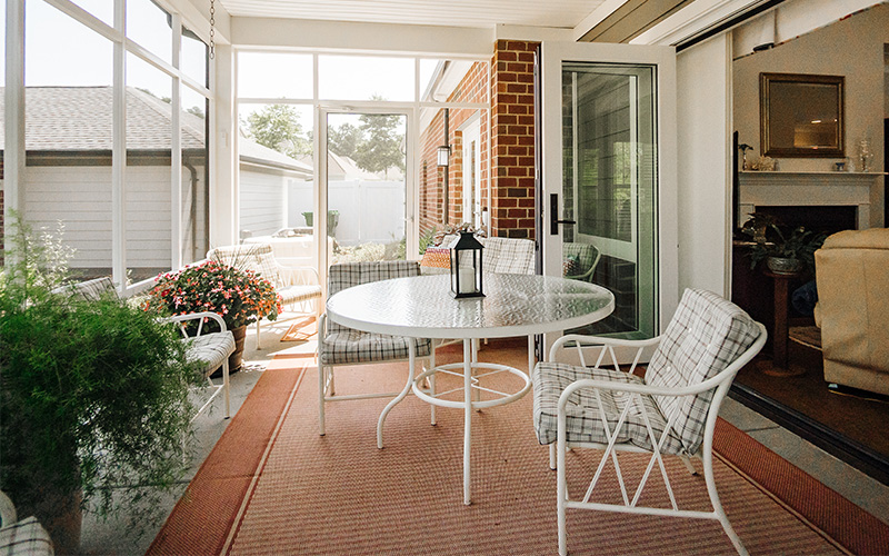 Bright sunroom with white patio furniture and potted plants in a senior living unit.