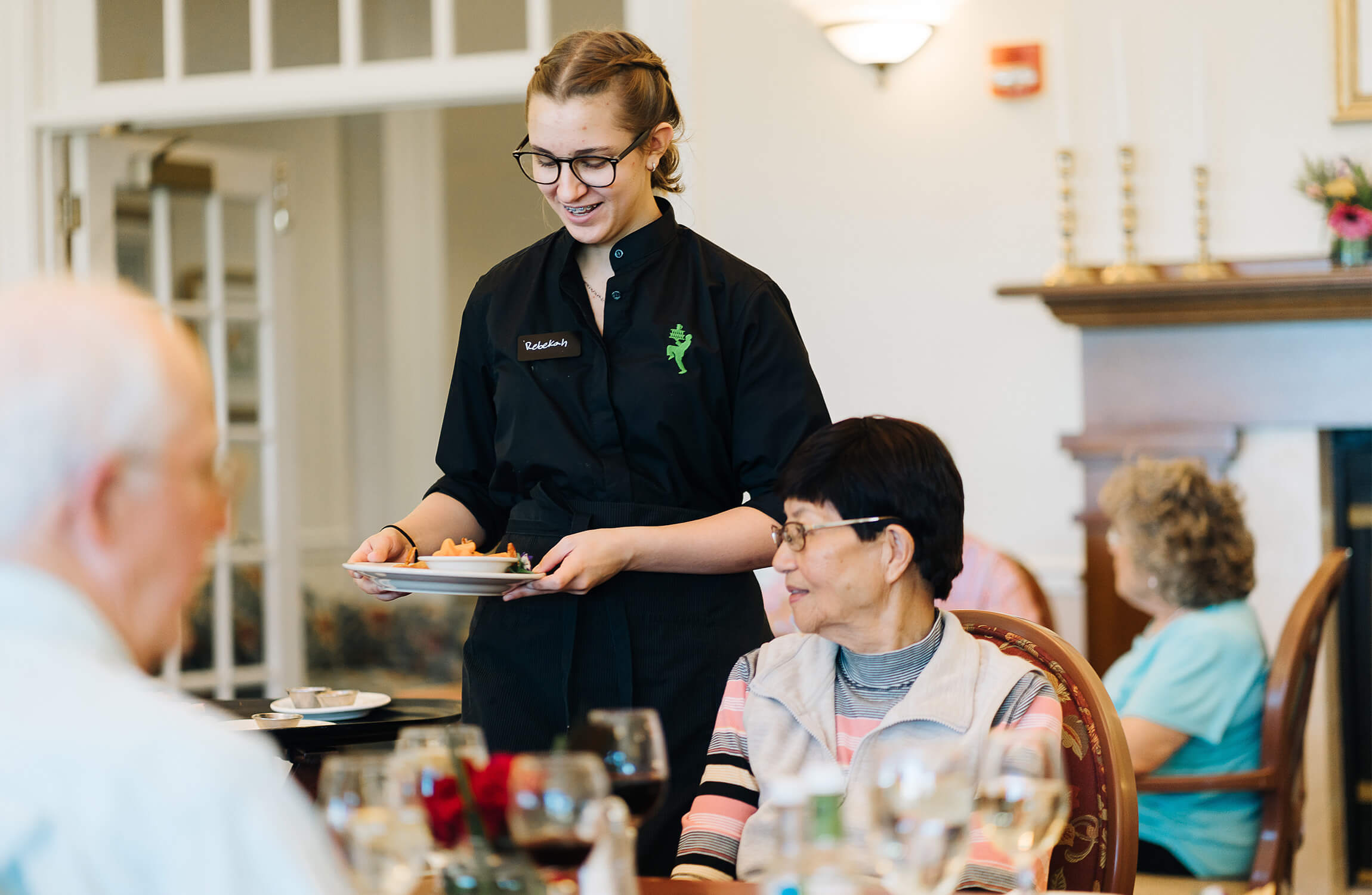 A staff member serves dinner to a seated senior woman in a communal dining area.