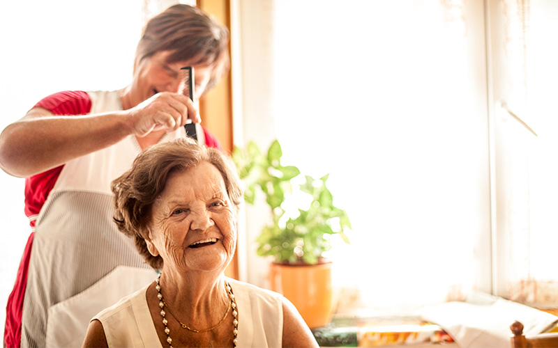 Elderly woman smiling as caregiver combs her hair in a bright room with a plant.