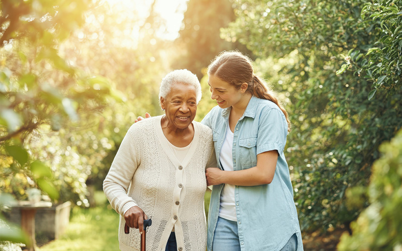 Elderly woman and caregiver smiling while walking in a sunlit garden.