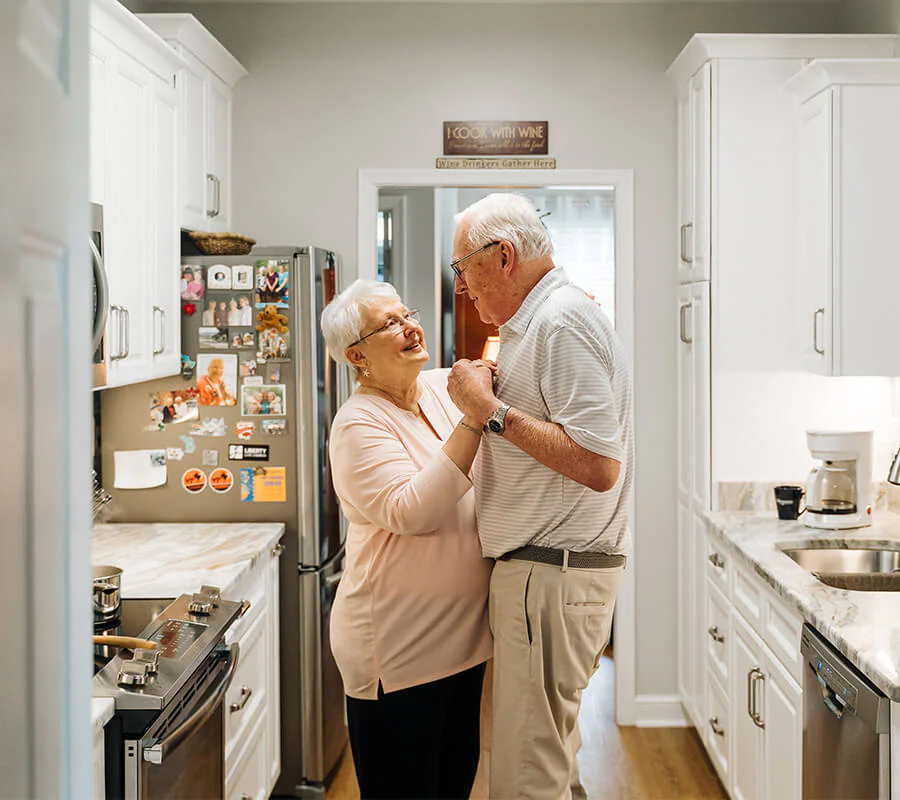 Elderly couple dancing in a cozy kitchen smiling warmly.