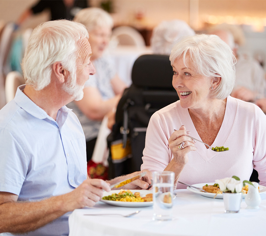 Elderly couple smiling while dining in a shared dining room of a senior community.