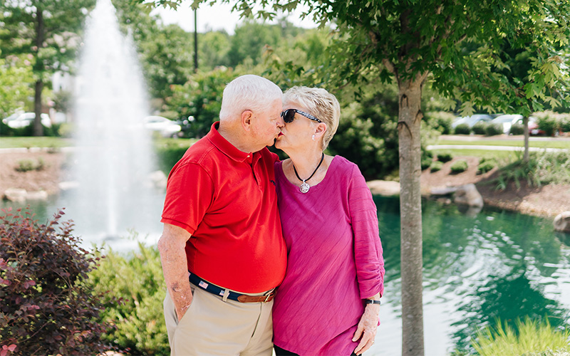 Elderly couple embracing near a pond with a fountain at a senior living community.