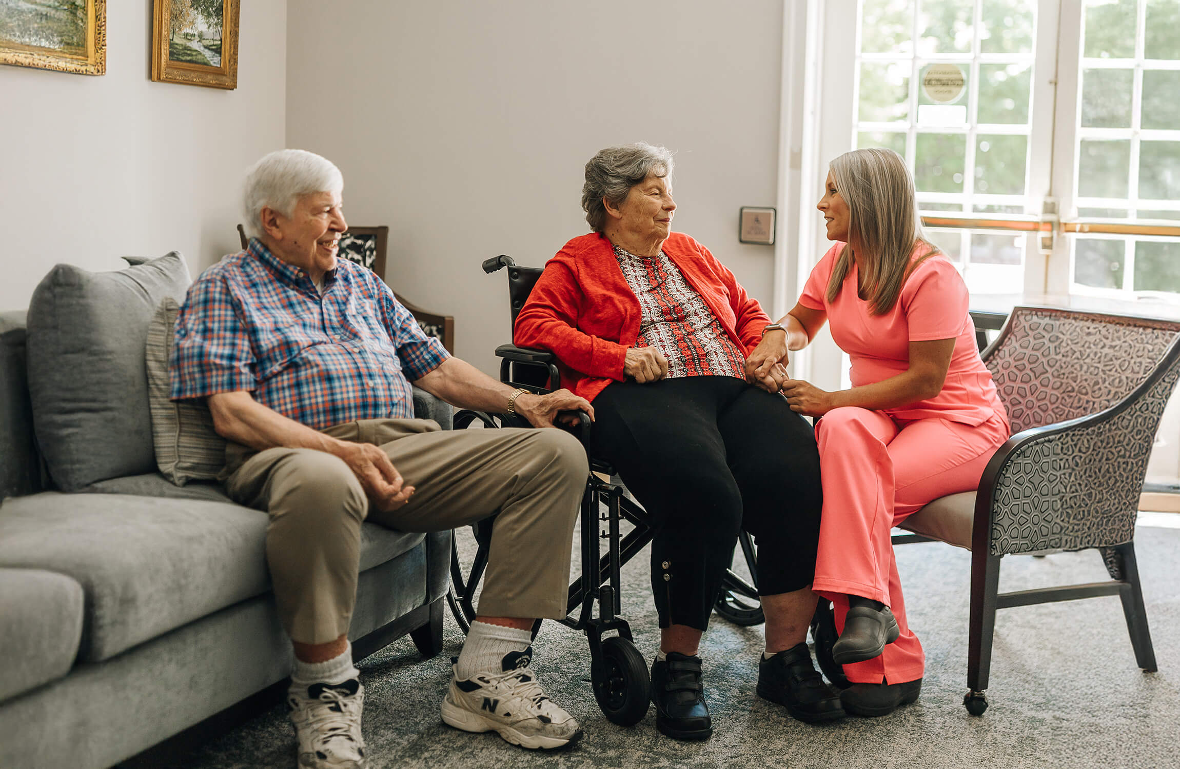 Elderly couple with a nurse in a bright communal living area holding hands and smiling.