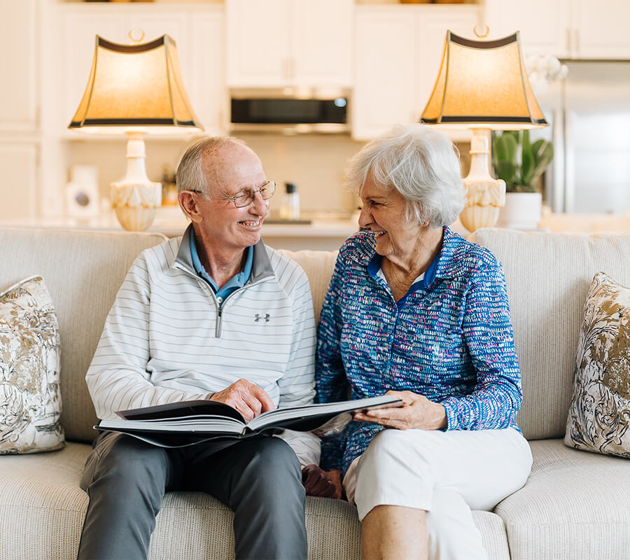 Elderly couple on sofa in cozy room, smiling while looking at a photo album.