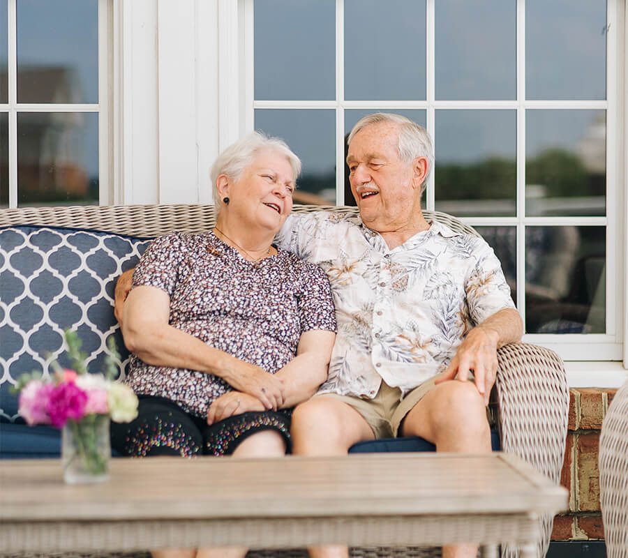 Smiling elderly couple sitting on a patio sofa, enjoying a sunny day together.