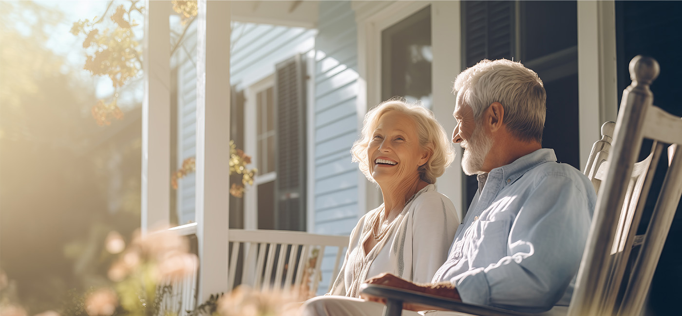Elderly couple smiling on the porch of a sunny senior living community unit.