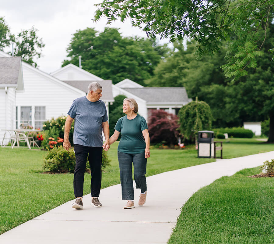 Elderly couple holding hands, walking on path with units and gardens in background.