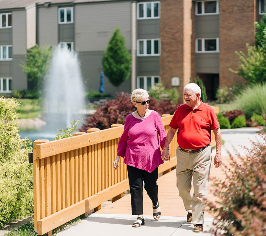 Elderly couple walking hand in hand on a sunny day by fountain in background.
