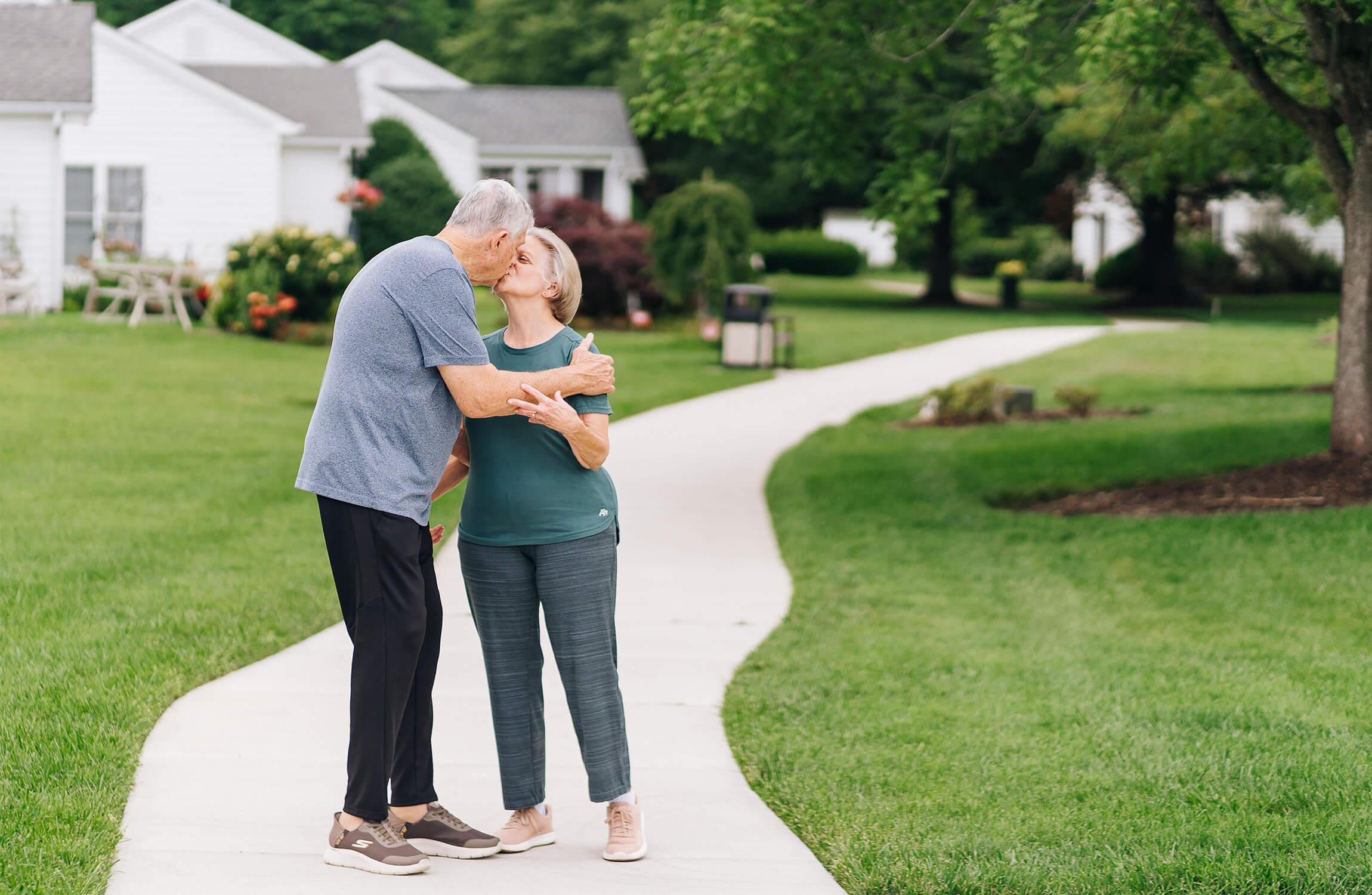 Elderly couple kissing on a walkway in a scenic senior living community.