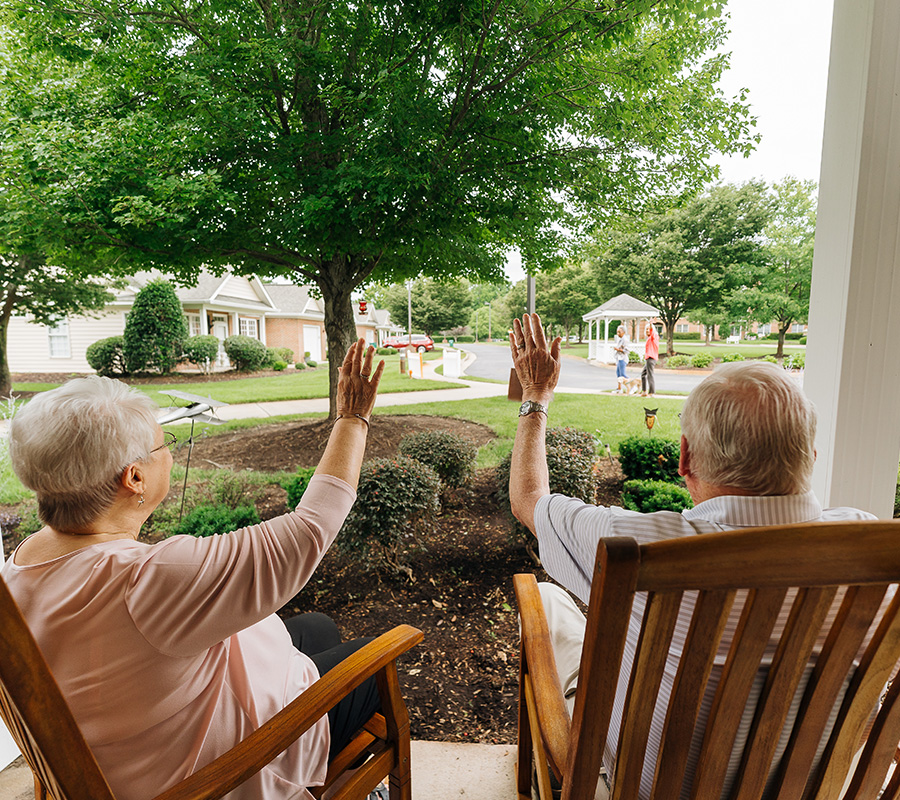 Elderly couple waving from porch at a community with lush greenery.