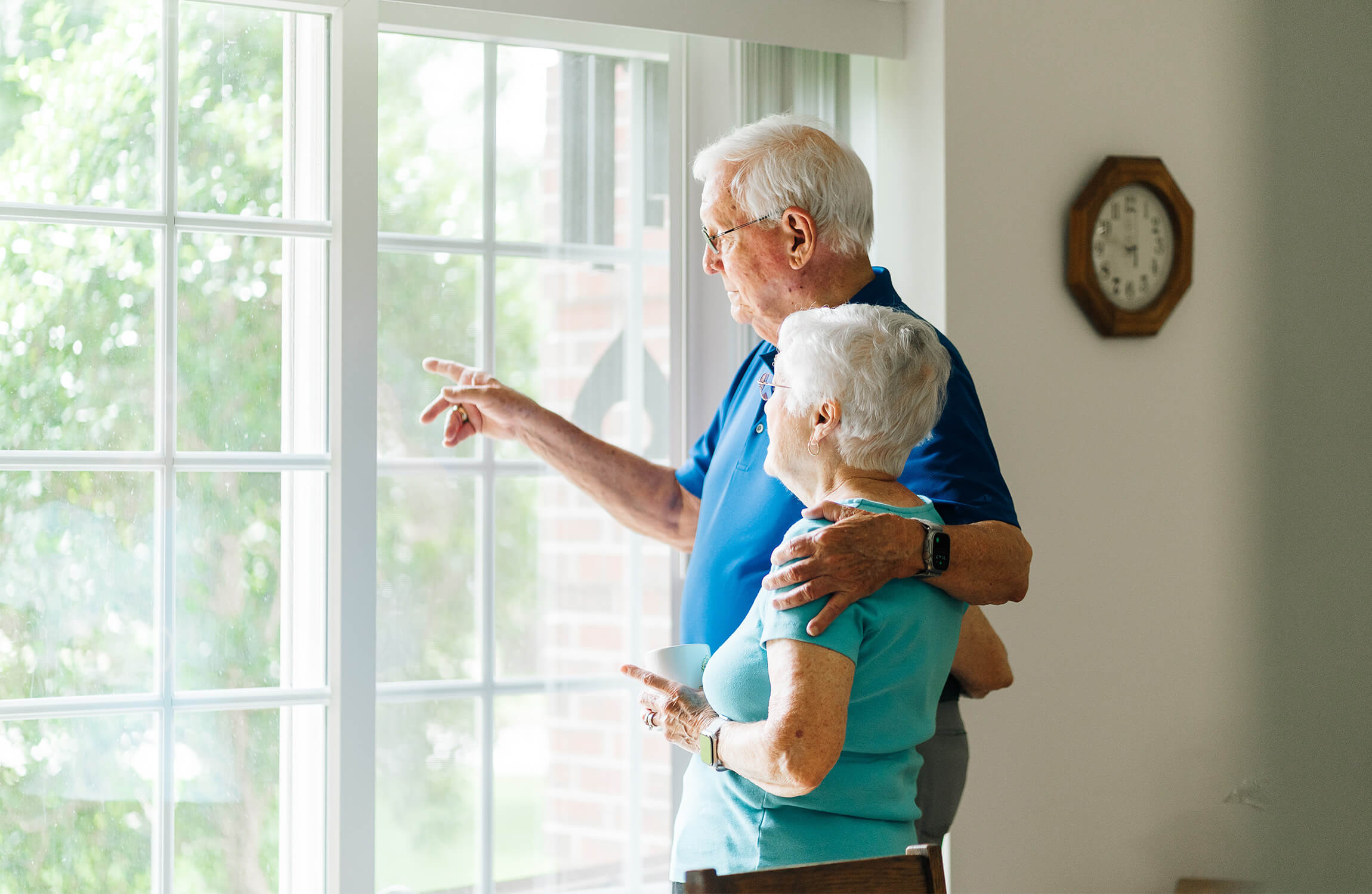 Elderly couple looking out the window in their unit with a clock on the wall