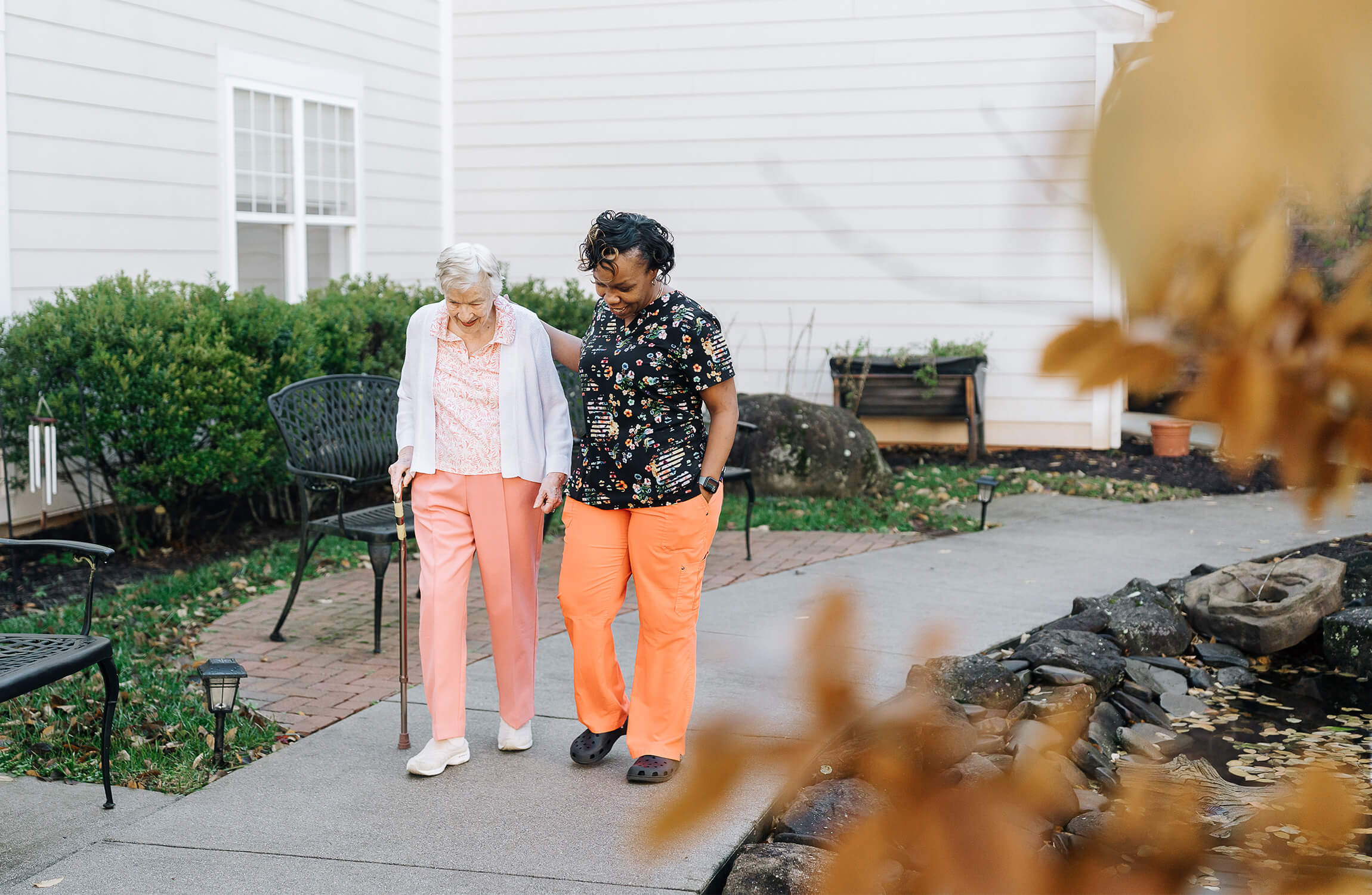 Elderly woman walking with caregiver in a senior community garden path.