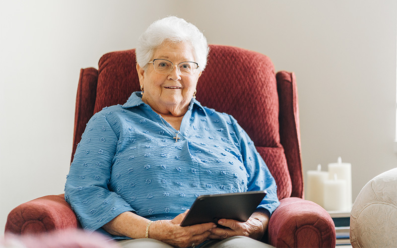 Elderly woman in blue shirt smiling while holding a tablet, seated on a red chair.