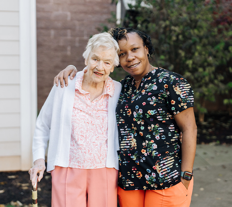 Elderly woman with caregiver outside in a senior living community garden.