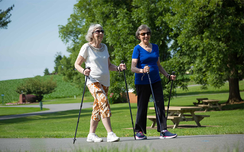 Two elderly women walking with poles in a green outdoor setting.