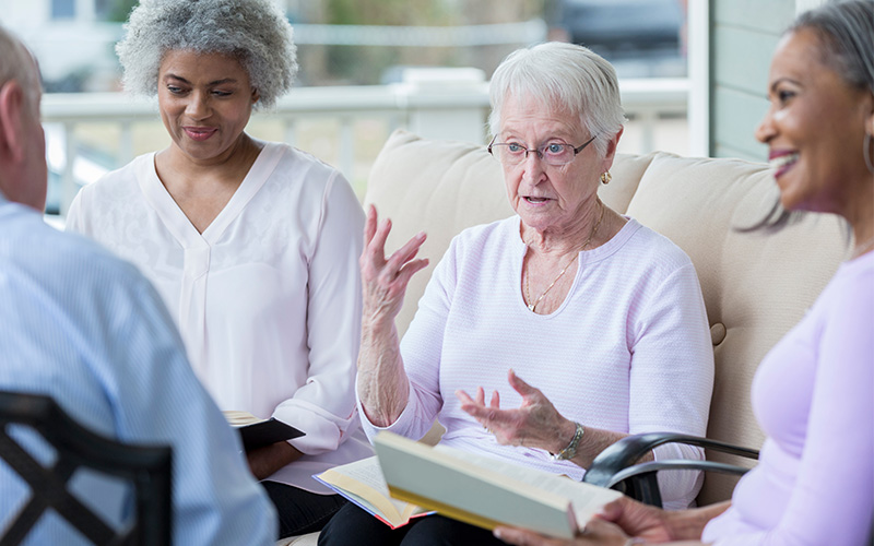 Elderly women engaged in a discussion while reading books together outdoors.