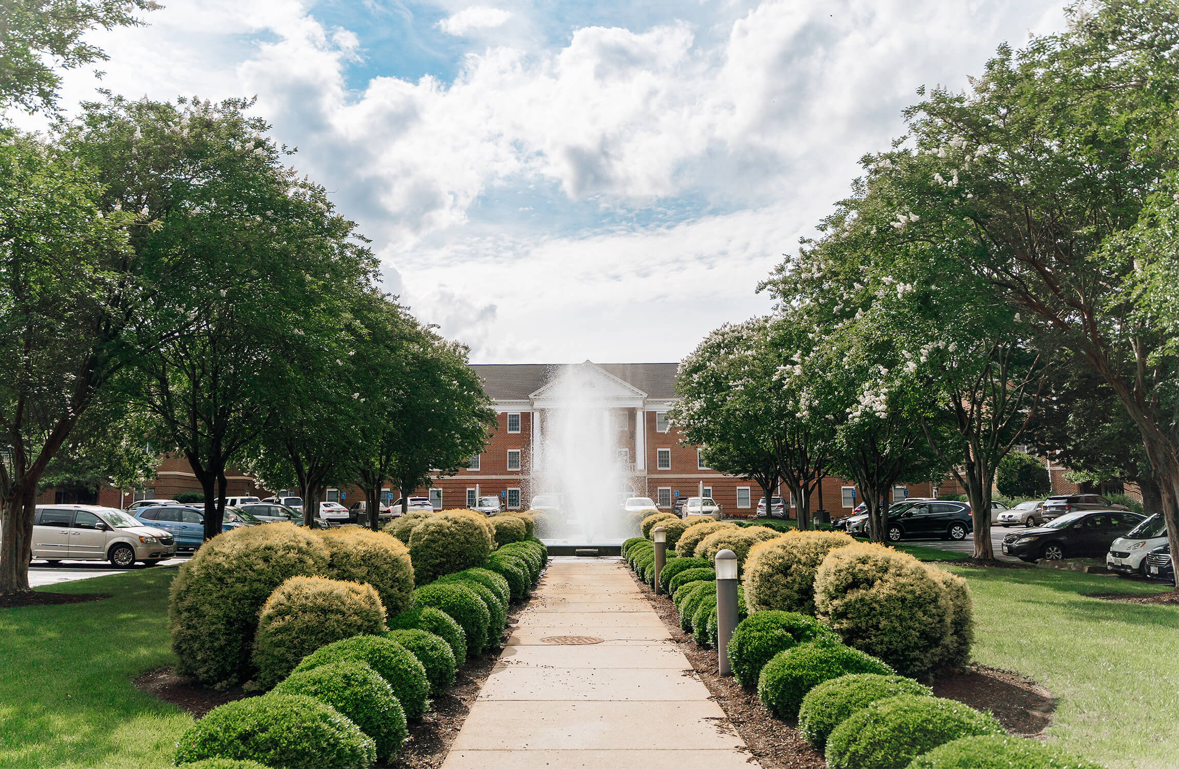 Pathway leading to a fountain surrounded by manicured bushes and trees, with a large building.