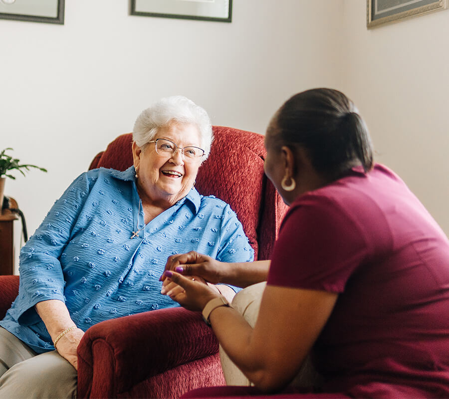 Elderly woman in blue chatting with caregiver in bright senior living unit.