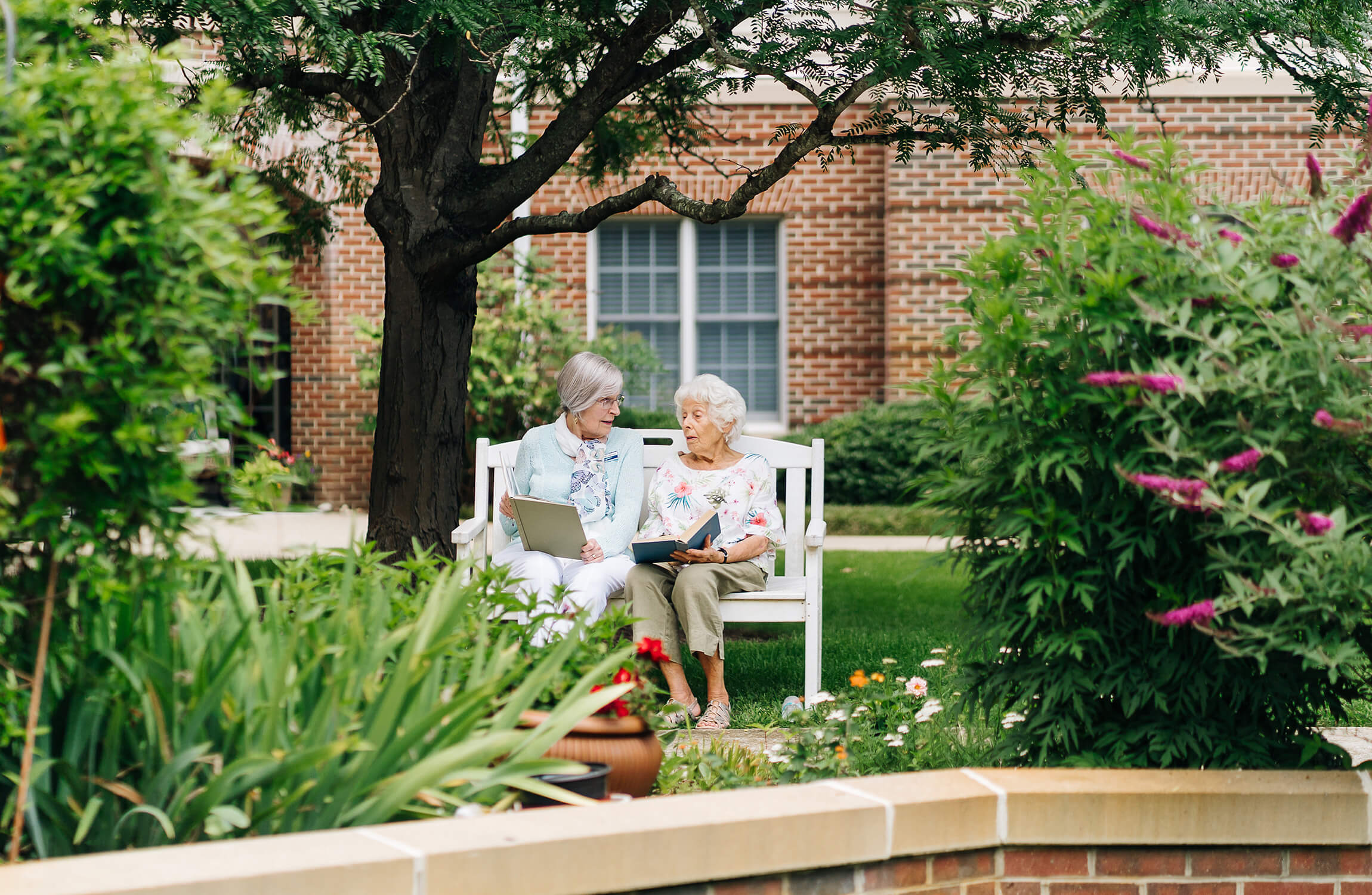 Two elderly women sitting on a garden bench sharing books under a tree.